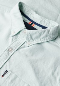 Camicia button-up verde menta chiaro con colletto strutturato, dotata di bottoni bianchi, texture morbida e colletto interno blu navy con strisce rosse e bianche.
