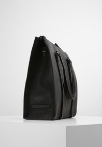 Zwarte leren totebag met een gestructureerde vorm, een gladde textuur, twee handvatten en een ritsluiting. Voorzien van een klein zwart logo plaatje.