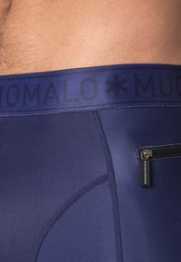 Navy blue swim trunks with a black elastic waistband featuring "MUCHACHO MALO" text. Includes a small zippered pocket on the side.