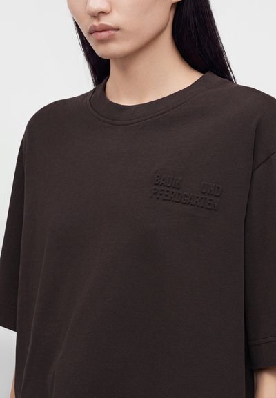 Femme portant un t-shirt marron foncé avec un texte en relief "Braum und Reehdgarten" sur le côté gauche de la poitrine, vue des épaules jusqu'au milieu du torse.