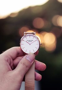 Orologio in oro rosa con quadrante bianco, indicatori delle ore sottili e bracciale a rete tenuto in mano contro uno sfondo sfocato.