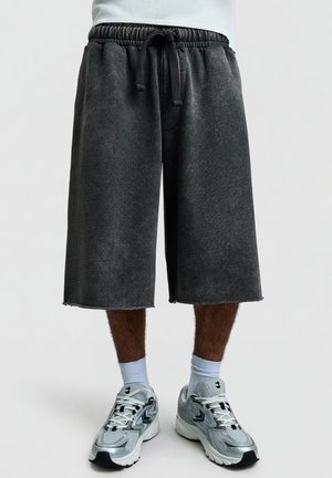 Egy férfi laza, térdig érő fekete rövidnadrágot visel, amelynek megkötőzsinórja van, fehér zoknit és ezüstfehér sportcipőt hord, és egyszerű háttér előtt áll.