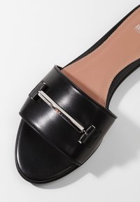 Sandale slide noire à bout ouvert avec large sangle en cuir et détail barre métallique argentée horizontale, semelle intérieure beige avec couture apparente.