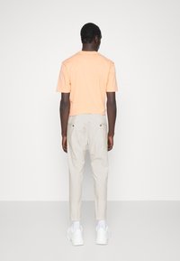 Kortärmad persiko färgad bomullströja i kombination med ljusbeige tapered byxor. Modellen bär vita sneakers och står framför en enhetlig bakgrund.