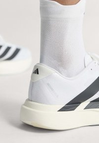 Nahaufnahme einer Person, die weiße Netzknöchelsocken und weiße Sportschuhe mit schwarzen Streifen auf hellem Hintergrund trägt.