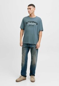 Grå T-shirt med korte ærmer og sort grafisk tryk; båret med lyse denim jeans og beige sko. Simpelt design, afslappet pasform.