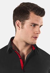 Chemise noire avec un col intérieur rouge et des détails de boutons, présentant un col pointu et une texture lisse, montrée en vue de profil.