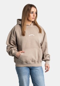 Sudadera beige de gran tamaño con un bolsillo frontal, capucha y texto impreso. Presenta puños y dobladillo acanalados, con una etiqueta de logo en la manga.