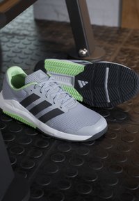 Paire de chaussures de course gris clair avec des bandes noires et des accents verts sur un sol de gym texturé noir près de l'équipement.