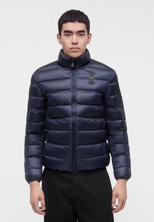 Giovane uomo che indossa una giacca imbottita blu scuro con collo alto e zip frontale, in piedi davanti a uno sfondo chiaro e uniforme.