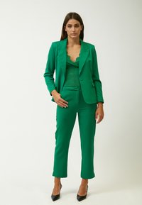 Groene op maat gemaakte kostuum met een getailleerd blazer en cropped broek, gecombineerd met een getextureerde kanten top. Geaccentueerd door zwarte puntige hakken.