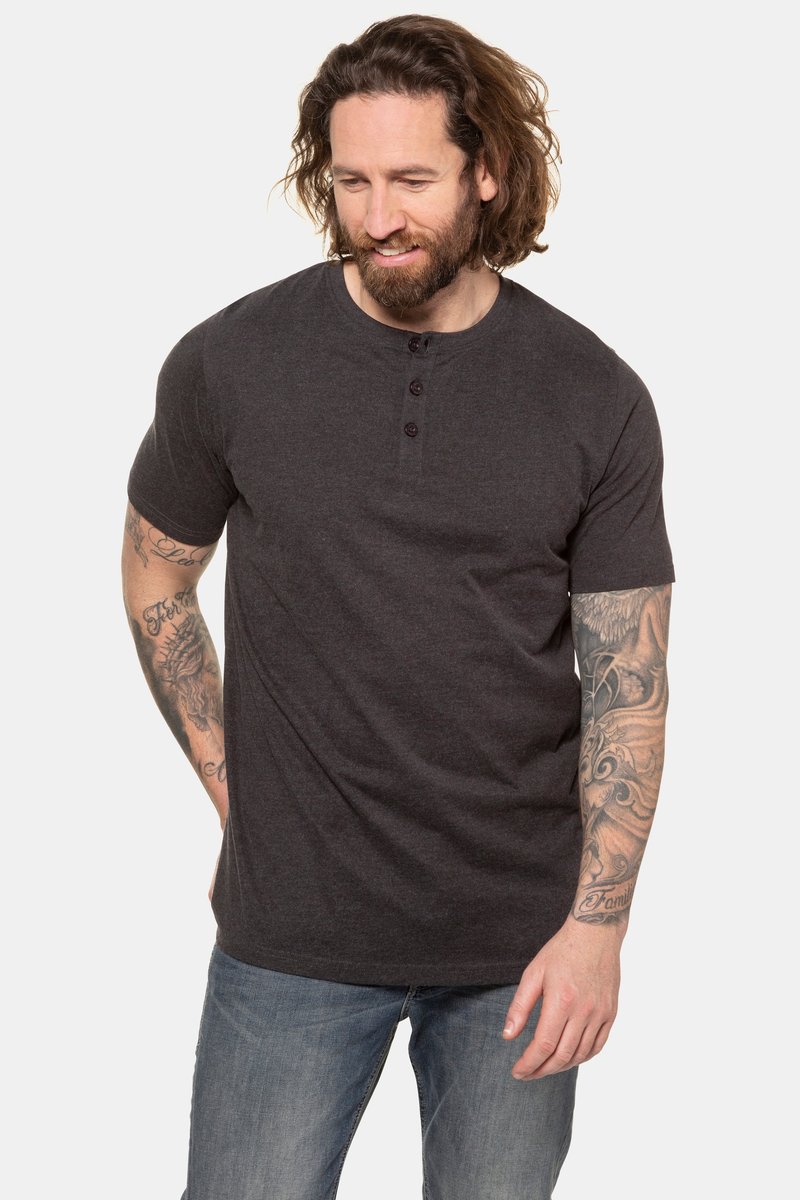 Mann mit langen Haaren und Tätowierungen an beiden Armen, der ein dunkelgraues Henley-Shirt und blaue Jeans trägt, schaut nach unten und lächelt.