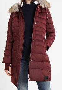 Kvinna i en lång vinröd vadderad kappa med luva i fuskpäls, marinblå tröja och blå jeans, med handen i fickan.