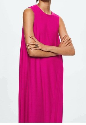 Femme portant une robe rose vif sans manches, bras croisés, avec une grande bague décorative à la main droite.