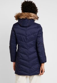 Tmavě modrý vatovaný kabát s kapucí lemovanou umělou kožešinou, vzor ve tvaru šipek, dvě boční kapsy a kontrastní hnědý lem. Střední délka s dlouhými rukávy.