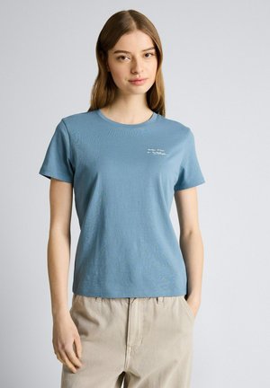 Młoda kobieta o prostych brązowych włosach, ubrana w niebieską koszulkę z małym białym napisem oraz beżowe spodnie, stojąca na tle jednolitego jasnego tła.