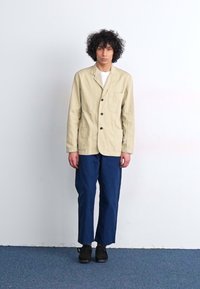Beige cotton-blend jacket with buttons and a chest pocket, paired with navy blue trousers and black shoes. Simple, relaxed fit.