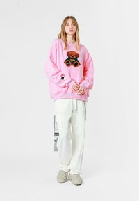 Sudadera rosa de gran tamaño con un diseño de oso en el frente, combinada con pantalones cargo blancos y zapatos ligeros de textura gris.