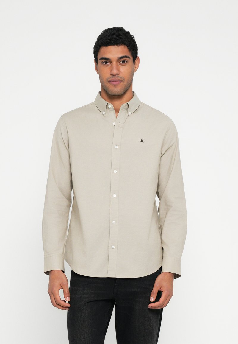 Camicia beige a maniche lunghe in tessuto di cotone, con colletto button-down e logo sul petto. Abbinata a jeans scuri.