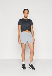 Μαύρο κοντομάνικο μπλουζάκι Nike με κόμπο μπροστά, συνδυασμένο με ανοιχτό γκρι σορτς και μαύρα αθλητικά παπούτσια. Λευκές κάλτσες με ορατό λογότυπο.