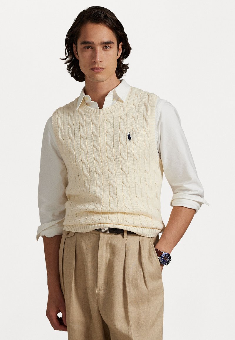 Suéter sin mangas de punto trenzado en crema sobre una camisa blanca con cuello, combinado con pantalones en beige claro. Presenta un pequeño logo en el pecho.