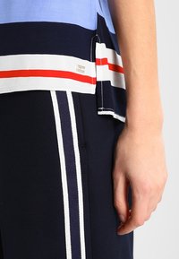 Casual top met horizontale blauwe, marineblauwe en witte strepen, voorzien van rode accenten en een logo-detail. Gepaard met marineblauwe broek met witte zijkanten.