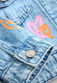 Mânecă de jachetă din denim albastru deschis, cu un nasture metalic gravat cu "ZÂMBETE FERICITE" și modele de inimioare roz și portocalii în apropiere.