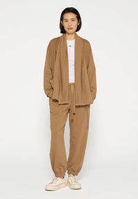 Giacca in maglia marrone chiaro con frontale aperto, abbinata a pantaloni joggers coordinati con cintura elastica. Sotto una t-shirt bianca e sneakers chiare.