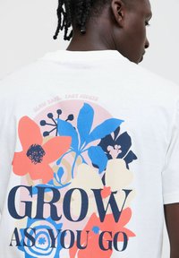 Hvit bomulls T-skjorte med et stort blomsterdesign i rødt, blått og mørke farger, med teksten "VOKS SOM DU GÅR" tydelig vist.