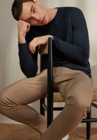 Långärmad marinblå skjorta, tan passande byxor, sittande på en stol med svart ram och flätad sits, lutande med huvudet på handen, trägolv.