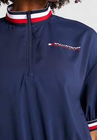 Marine sports polo med halv lynlås krave, med en stribet rød og hvid bånd, og "TOMMY SPORT" logoet i hvid. Glat stof tekstur.