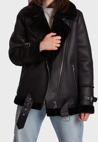 Svart läderjacka med fårskinnskrage, silverdragkedjor, justerbara remmar och en strukturerad yta. Bärs över en grå topp och blå jeans.