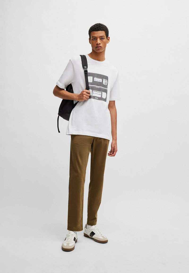T-shirt en coton blanc avec impression graphique, associée à un pantalon vert olive. Le modèle porte un sac à dos noir et des baskets blanches et marron.