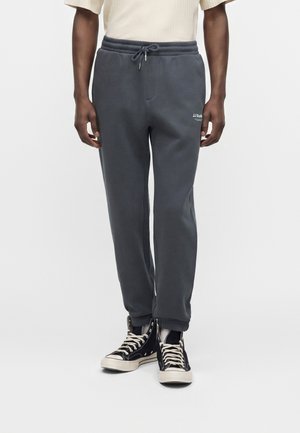 Pantalon de survêtement gris foncé avec taille à cordon de serrage et petit logo blanc au-dessus de la cuisse gauche, porté avec des baskets montantes noires et lacets blancs.