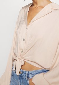 Lähikuva henkilöstä, joka on pukeutunut vaaleanpunaiseen, kaulukselliseen sitomiseen paitaan, joka on yhdistetty korkeavyötäröisiin sinisiin farkkushortseihin. Vatsan alue näkyy.