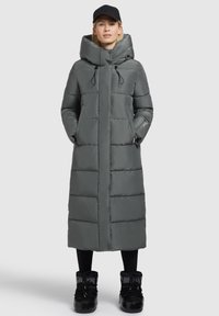 Lange, grijze puffercoat met capuchon, voorzien van een quilted ontwerp, voorzakken en toggelkoordjes; gecombineerd met zwarte laarzen.