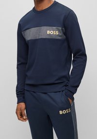 Marinblå sweatshirt med grå horisontell rand och guld "BOSS"-logotyp på bröstet. Mjuk kvalitet, långa ärmar, ribbade muddar och fåll.