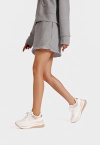 Sneaker bianchi con accenti beige, caratterizzati da una texture liscia, design con lacci e una suola in gomma spessa per supporto. Outfit casual grigio.