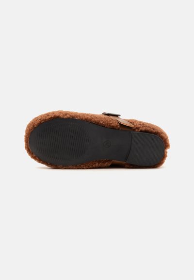 Chausson marron duveteux vu par le dessous avec une semelle en caoutchouc noir et une bride à boucle sur fond blanc.
