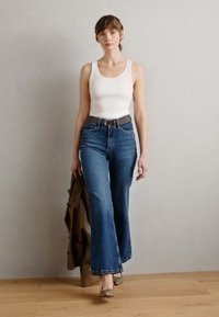 Bílý tílko, vysoce pasované modré džíny s rozšířenými nohavicemi, hnědý opasek a béžové podpatky. Modelka drží hnědou bundu. Světlé pozadí.