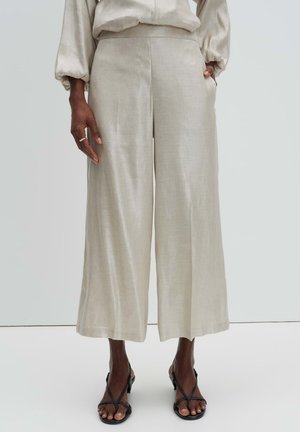 Vrouw draagt beige wijde cropped broek met een beige blouse met lange mouwen en zwarte sandalen met hoge hak en bandjes, staand tegen een effen achtergrond.