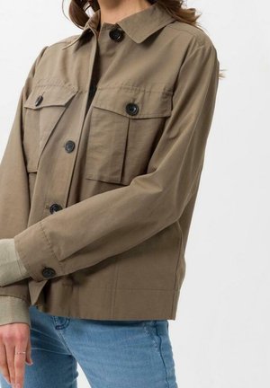 Nainen, joka käyttää vaaleanruskeaa, napitettavaa takkia, jossa on läppätaskut rinnassa ja ribbineuloksiset hihansuut, yhdistettynä sinisiin farkkuihin, yksiväristä taustaa vasten.