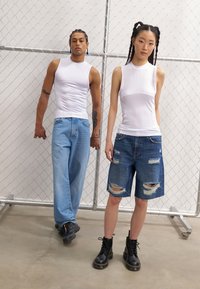 Hvit tettsittende ermeløs topp, lys blå baggy jeans, mørkeblå ripped shorts og svarte snørehæler, mot en bakgrunn av kjede-nett.
