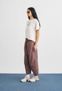 Μπεζ t-shirt με γραφική λεπτομέρεια, συνδυασμένο με φαρδιά, καφέ φόρμα. Το μοντέλο φορά ασημένια γυαλιά ηλίου και ανοιχτά αθλητικά παπούτσια.