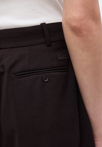 Gros plan sur une personne portant un pantalon noir avec une poche arrière boutonnée et un petit logo crocodile brodé sur la ceinture.