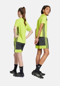 Camiseta y pantalones cortos deportivos de un verde lima brillante con rayas negras, combinados con zapatos de fútbol y calcetines negros. Textura suave, diseño deportivo.