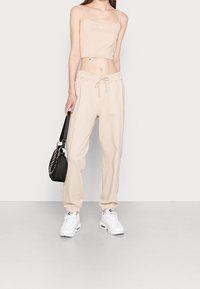 Béžový crop top a odpovídající joggers vyrobené z měkké tkaniny, s elastickým pasem. Bílé tenisky a malá černá taška s řetízkovým popruhem.