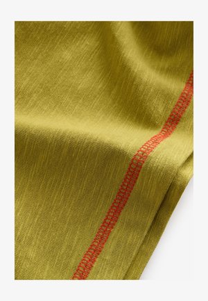 Zelená látka se strukturovaným povrchem a červeným dekorativním stehem podél jednoho okraje. Materiál působí měkce a lehce.