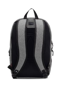 Mochila de tela gris con correas de malla negra, un asa en la parte superior y un parche con logo. Presenta un diseño estructurado y bolsillos con cremallera a los lados.