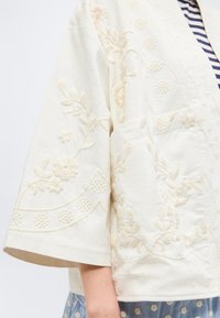 Veste couleur crème avec broderie florale, manches larges trois-quarts et une poche avant, portée sur un haut rayé et une jupe bleue à pois.